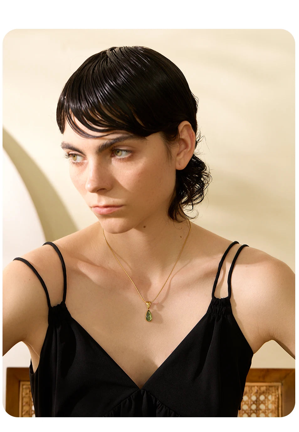 Mulher com cabelo curto e molhado, usando vestido preto e colar dourado com pingente verde.