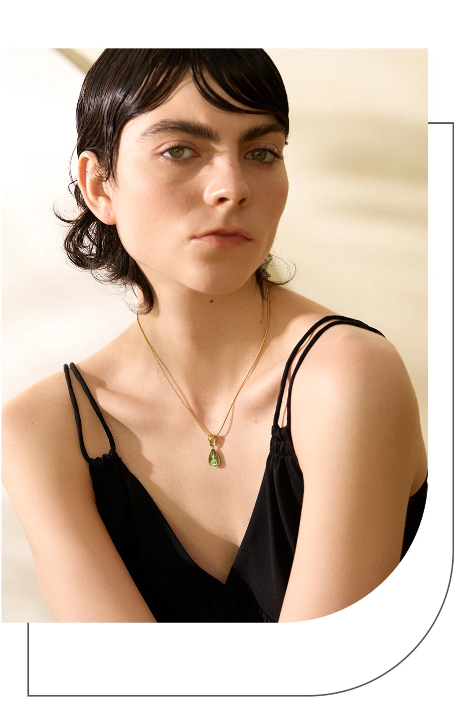 Mulher de cabelos curtos usando vestido preto e colar dourado com pingente verde.