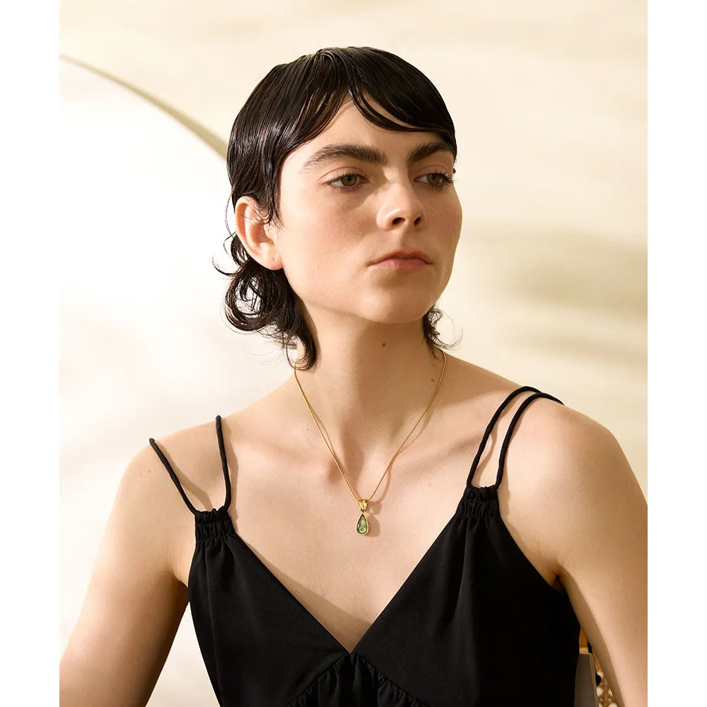 Mulher com cabelo curto usa vestido preto e colar dourado com pingente verde, fundo claro.