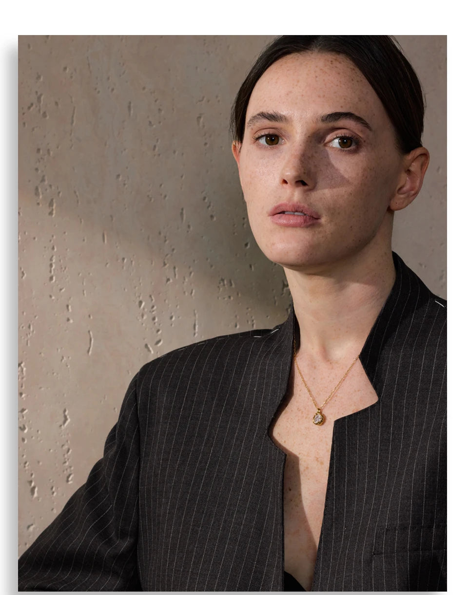 Mulher usando blazer risca de giz escuro e colar dourado, fundo bege texturizado.