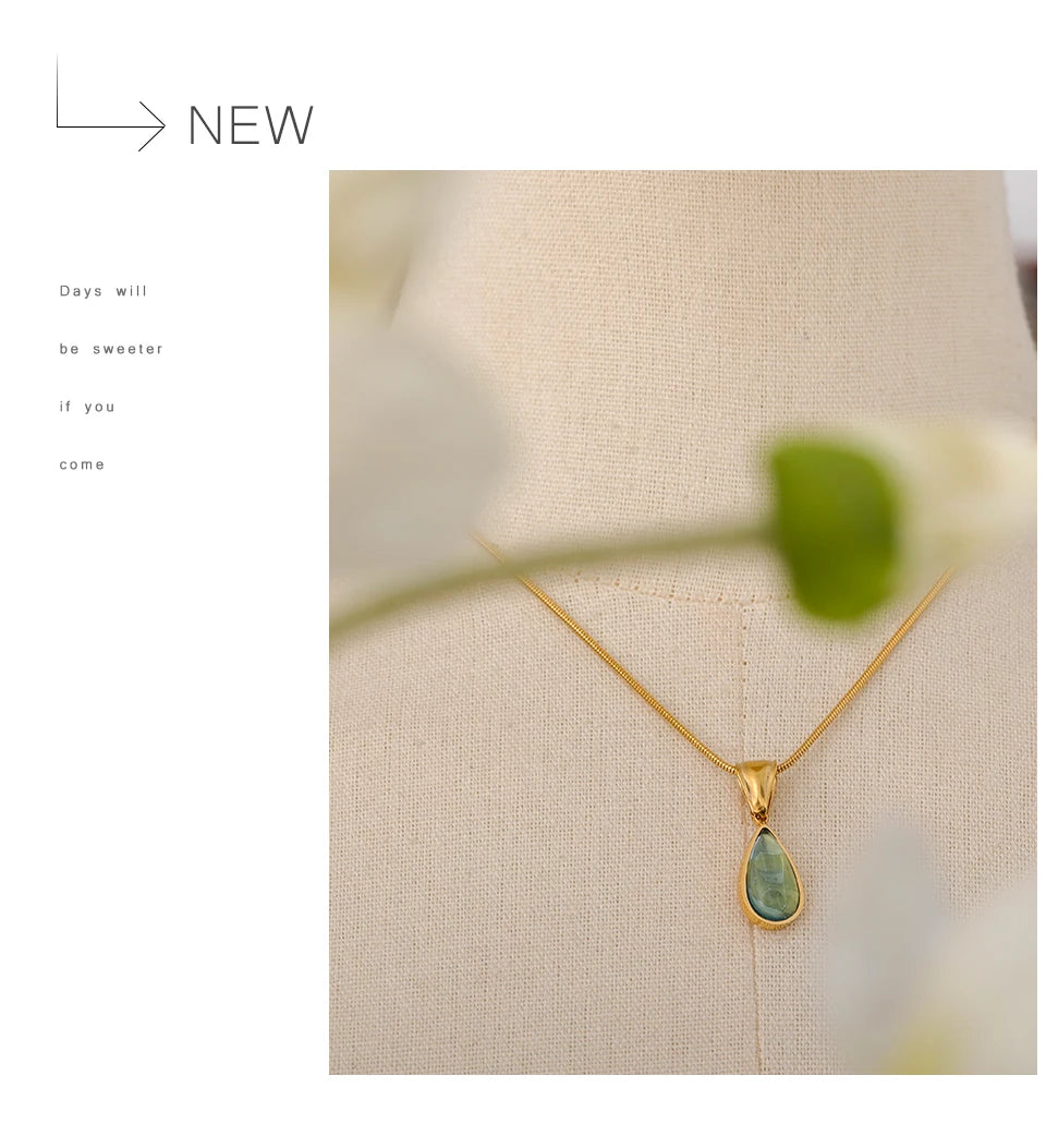 Colar dourado com pingente de pedra verde em busto de tecido, flores desfocadas na frente.