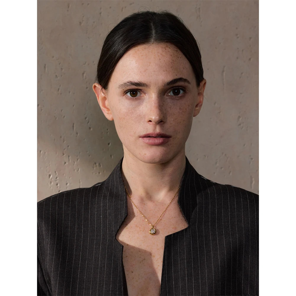 Mulher com colar dourado de pingente, blazer risca de giz, fundo bege texturizado